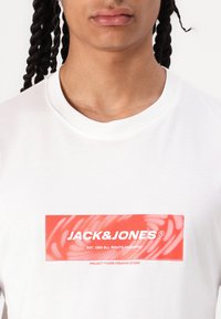Hvid bomulds T-shirt med et centralt rødt graphic med teksten "JACK&JONES" og et svungent mønster, rund halsudskæring, ingen synlige metaldele.