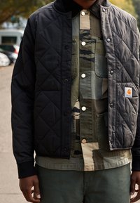 Zwarte gewatteerde jas met drukknopen over een overhemd met camoufleringspatroon. Warme stof, contrasterende texturen en zichtbare patch met logo.