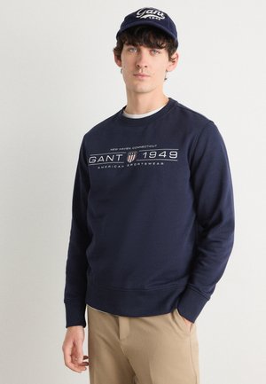 Navy sweatshirt with round neck, ribbed cuffs, and hem. Features white text graphic; worn with khaki trousers and a navy cap.