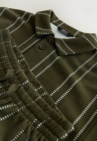 Olivgrünes Poloshirt mit weißen vertikalen Streifen, gerippter Textur und zwei braunen Knöpfen am Kragen. "NEXT"-Etikett sichtbar.