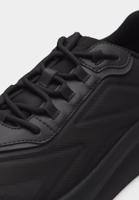 Κοντινή εικόνα μαύρου αθλητικού παπουτσιού με υφασμάτινη υφή, συνθετικές επενδύσεις και μοναδικό σύστημα δεσίματος με θηλιές σε λευκό φόντο.