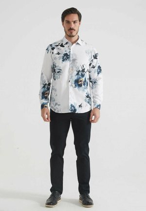 Chemise blanche à boutons avec un motif floral bleu foncé, manches longues et col classique, associée à un pantalon noir et des chaussures noires.