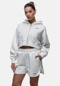 Felpa bianca corta e pantaloni abbinati realizzati in un tessuto morbido. La felpa presenta un logo blu e una zip frontale; i pantaloni hanno un elastico in vita.