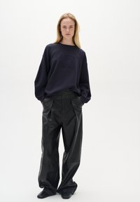 Námořnická mikina s Embosovaným logem, spárovaná s volnými černými koženými kalhotami. Modelka pózuje v minimalistickém stylu na neutrálním pozadí.