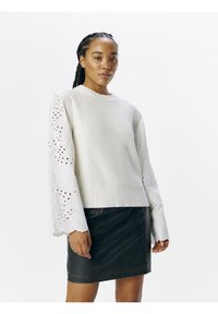 Maglione bianco a maglia con orlo a costine e maniche in pizzo ad occhiello dettagliato, abbinato a una gonna di jeans nera, creando un look stratificato e tessurizzato.