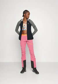 Giacca con zip nera e grigia indossata sopra un top corto colorato, abbinata a pantaloni rosa con dettagli neri e tasca con cerniera.