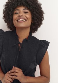 Rochie neagră cu corset plisat, mâneci volănaș și închidere cu fundă la gât. Textură netedă cu un design elegant și rafinat.