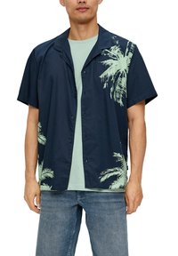 Korte mouwen, marineblauwe knoopsluiting shirt met lichtgroene palmbomenprint, gecombineerd met een mintgroen t-shirt, gedragen met blauwe jeans.