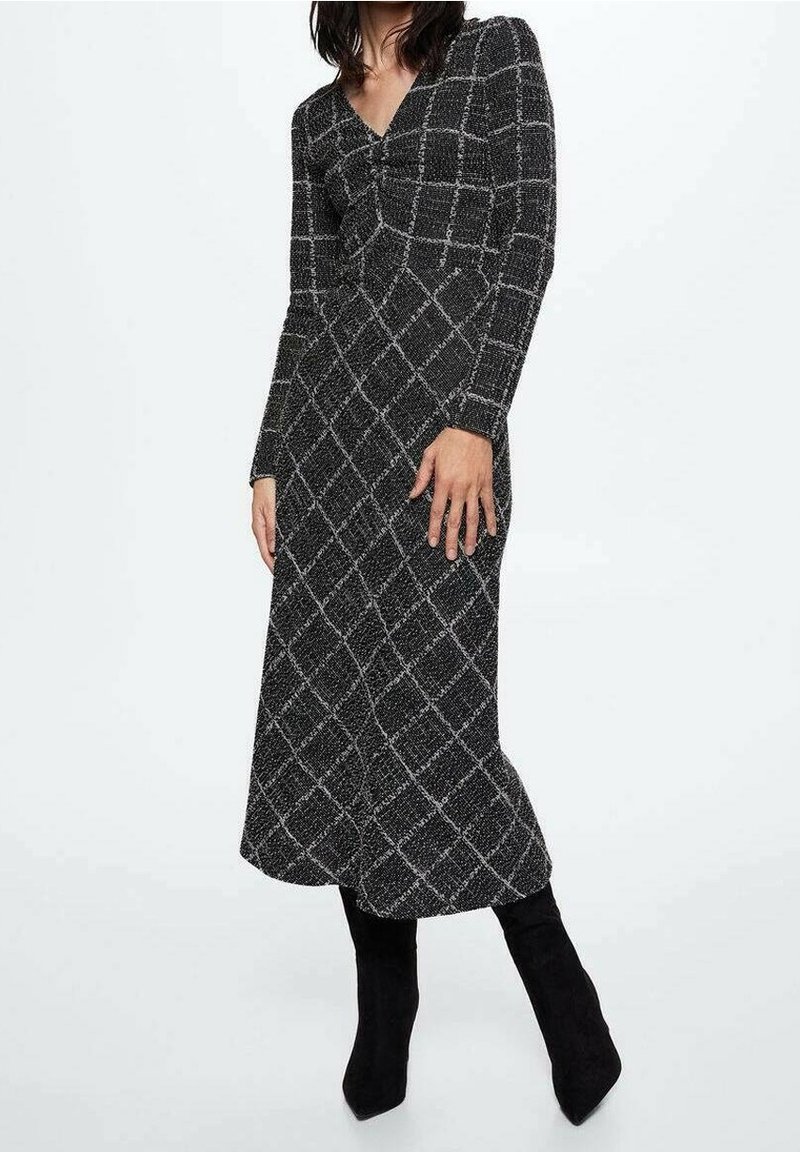 Femme portant une robe midi à manches longues noire et blanche à carreaux, associée à des bottes noires à talons, sur un fond blanc.