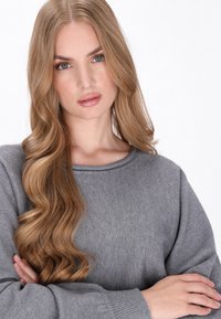 Pulover gri tricotat cu guler rotund și mâneci lungi, ușor supradimensionate. Textură netedă și croială lejeră, purtat de o persoană cu păr lung.