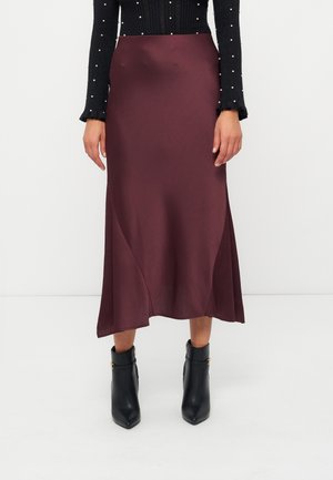 Femme portant une jupe midi violet foncé, un haut à manches longues noir orné de perles et des bottines noires, debout sur un fond blanc.