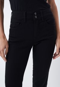 Gros plan d'une personne portant un jean noir taille haute avec fermeture à double bouton et poches avant, mains détendues sur les côtés.
