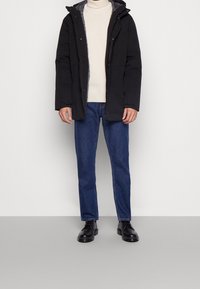 Chaqueta acolchada negra con capucha, forro gris, cierre frontal a presión, combinada con un suéter crema, jeans azules y botas negras.