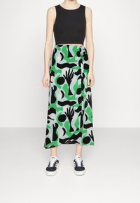 Zwarte mouwloze crop top gecombineerd met een maxi rok in groen en zwart met abstracte vormen en handmotieven, gedragen met donkere sneakers.