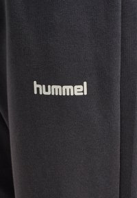 Ciemnoszara tkanina z białym logo "hummel" nadrukowanym na materiale, ukazująca szczegółową fakturę i przeszycia.