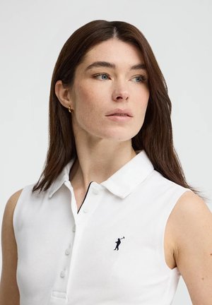 Vrouw met middellang bruin haar, die gouden oorringen draagt en een mouwloze witte blouse met kraag en een klein donker logo op de borst.