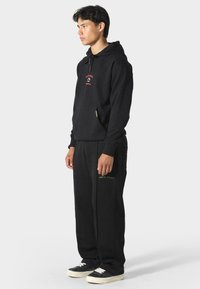 Sudadera de algodón negra con bolsillo frontal y cordones, combinada con pantalones de chándal negros con texto dorado. Zapatillas de suela blanca completan el look.