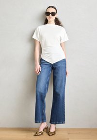 Top blanco de manga corta con cintura fruncida, combinado con vaqueros de tiro alto y pierna ancha en azul y mules de tacón con estampado de leopardo amarillo.