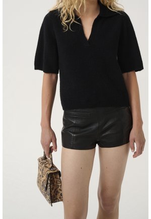 Woman wearing black short-sleeve sweater and black leather shorts, holding a small leopard-print handbag by her side.