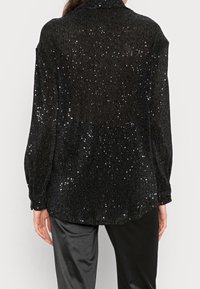 Black patterned jacket with ribbed texture, adorned with shiny sequins, featuring a collar and long sleeves, paired with dark trousers.