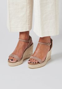 Sandalias de cuña de ante beige con dos correas frontales y una correa en el tobillo, con una plantilla de yute trenzado y suela texturizada, adecuadas para el uso casual.