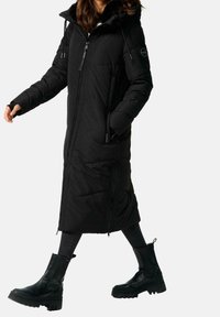 Sort lang dunjakke med pelsforet hætte, lynlåslukning, to sidelommer og quiltet mønster. Kombineret med sorte ankelstøvler.