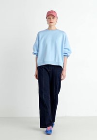 Sweatshirt oversize azul claro com mangas bufantes, jeans de perna larga azul escuro e um boné rosa. Tênis azuis com cadarços rosas estão usados.