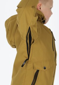 Braune wasserdichte Jacke mit Kapuze, schwarzem Reißverschluss, seitlicher Belüftung und einer Fronttasche mit Druckknopf. Glatte Textur und modernes Design.