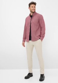 Uomo sorridente, che indossa una camicia a quadri rossa sopra una maglia nera, pantaloni beije e stivali stringati neri, in piedi contro uno sfondo bianco.