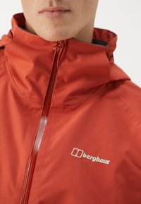Czerwona wodoodporna kurtka z wysokim kołnierzem, pełnym zamkiem błyskawicznym i białym logo Berghaus. Gładka tekstura, nowoczesny design.