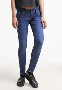 Jean ajustados en denim azul oscuro, con un clásico diseño de cinco bolsillos y costuras sutiles. Combinados con zapatos negros perforados.
