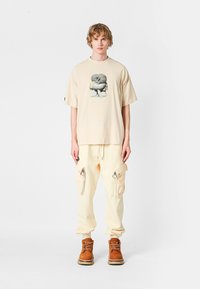 Camiseta beige de gran tamaño con estampado gráfico, combinada con pantalones cargo amarillos claros que tienen cremalleras y acentos de anilla. Botas marrones con cordones.