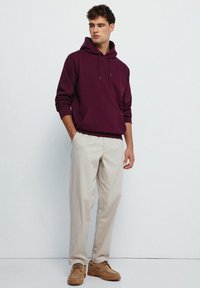 Felpa bordeaux con tasca frontale e cappuccio con coulisse, abbinata a pantaloni beige chiaro e scarpe brown con lacci. Tessuto liscio in tutto.