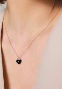 Złoty naszyjnik z łańcuchem, z małym czarnym zawieszeniem w kształcie serca, ozdobionym drobnym diamentem, spoczywającym na nagiej szyi.