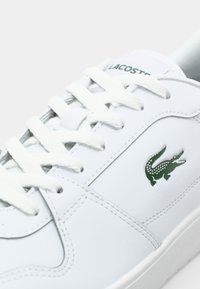 Witte leren sneaker met groen Lacoste-logo, platte veters, ronde neus en gestikte details op de bovenkant. Glad oppervlak en minimalistisch ontwerp.