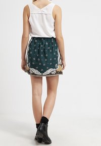 Groene rok met bloemen gouden accenten en witte randen; elastische tailleband; gedragen met een witte mouwloze top en zwarte enkellaarzen.