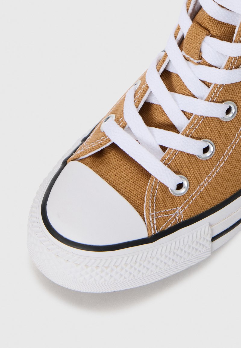 Tan canvas-sneakers med vita snören, en gummitå och en texturerad vit sula med svart kantdetalj.