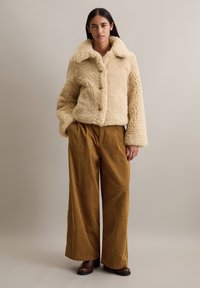 Veste en fausse fourrure beige avec un large col et une fermeture à boutons à l'avant, assortie à un pantalon large en velours côtelé marron taille haute et des bottes marron.