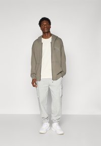 Grå zip-up hættetrøje med lomme, beige T-shirt, grå cargo sweatpants og hvide sneakers. Afslappet outfit med løs pasform og enkelt design.