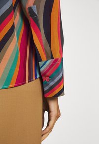 Personne portant une chemise à manches longues avec des rayures verticales multicolores et des poignets boutonnés, associée à une jupe ou un pantalon beige.