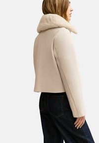 Beżowa krótka kurtka z miękkim kremowym kołnierzem, gładkim materiałem, prostym krojem i minimalistycznym designem. W zestawieniu z ciemnymi spodniami dżinsowymi.