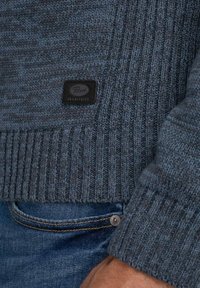 Jersey de punto azul oscuro con bajo acanalado, que presenta un patrón texturizado y un pequeño parche de cuero negro etiquetado como "Parrot Industries."