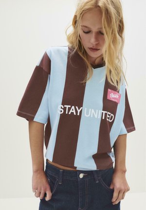 Jeune femme aux cheveux blonds regardant vers le bas, portant une chemise à rayures brunes et bleu clair avec le texte "RRESTEZ UNIS" et un jean bleu foncé.