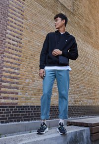 Sort hoodie med snore, lyseblå bukser med oprullede manchetter og sorte sneakers med grønne accenter, sat op mod en tekstureret mur.