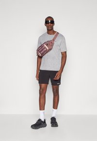 Σακίδιο ώμου σε μπορντό χρώμα με αντίθετους ιμάντες· γκρι μπλούζα, μαύρα σορτς και μαύρα sneakers· μοντέλο που φοράει υπερμεγέθη γυαλιά ηλίου.