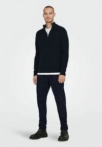Navy pullover with a half-zip collar, made of textured fabric. Worn over a white shirt, paired with navy pants and black shoes.