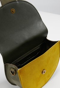Mała torebka na ramię z otwartą klapką, ciemnozielonym skórzanym wnętrzem i musztardowo-żółtą zamszową kieszenią na zewnątrz.