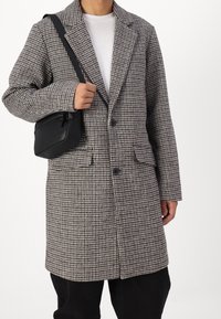Abrigo con patrón de pata de gallo en tela beige y negra, con cuello de solapa con muesca, dos bolsillos delanteros y cierre de un solo botón.