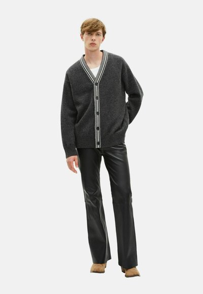 Cardigan in lana grigio carbone con scollo a V, bordo a righe bianche e bottoni. Indossato con pantaloni in pelle nera aderenti e scarpe beige.