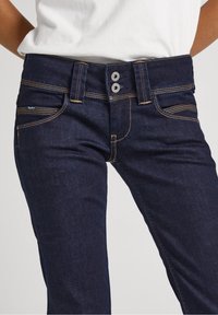 Pantalones vaqueros de mezclilla azul oscuro con corte slim, que presentan costuras en amarillo en contraste, cierre de dos botones y bolsillos delanteros con ligero detalle.